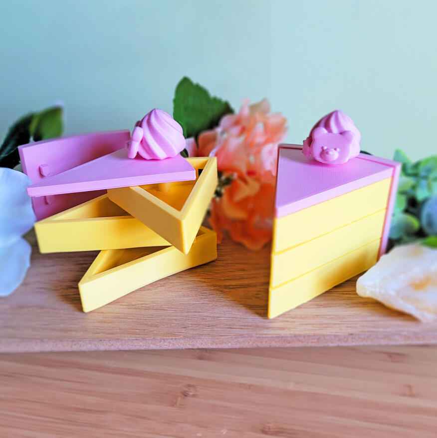 Untitled Two pink and yellow 3D printed boxes, modeled to look like slices of cake. The boxes have 3 compartments, each one a "layer" of cake. There is a small decoration on the top of them. One of them is closed, and one of them is open, showing how the compartmen